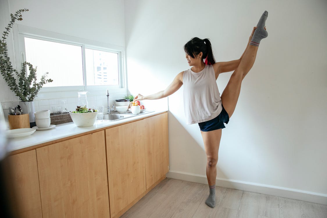 Full length of young female in sleepwear standing in kitchen and doing splits with leg up near counter while making tea for meal