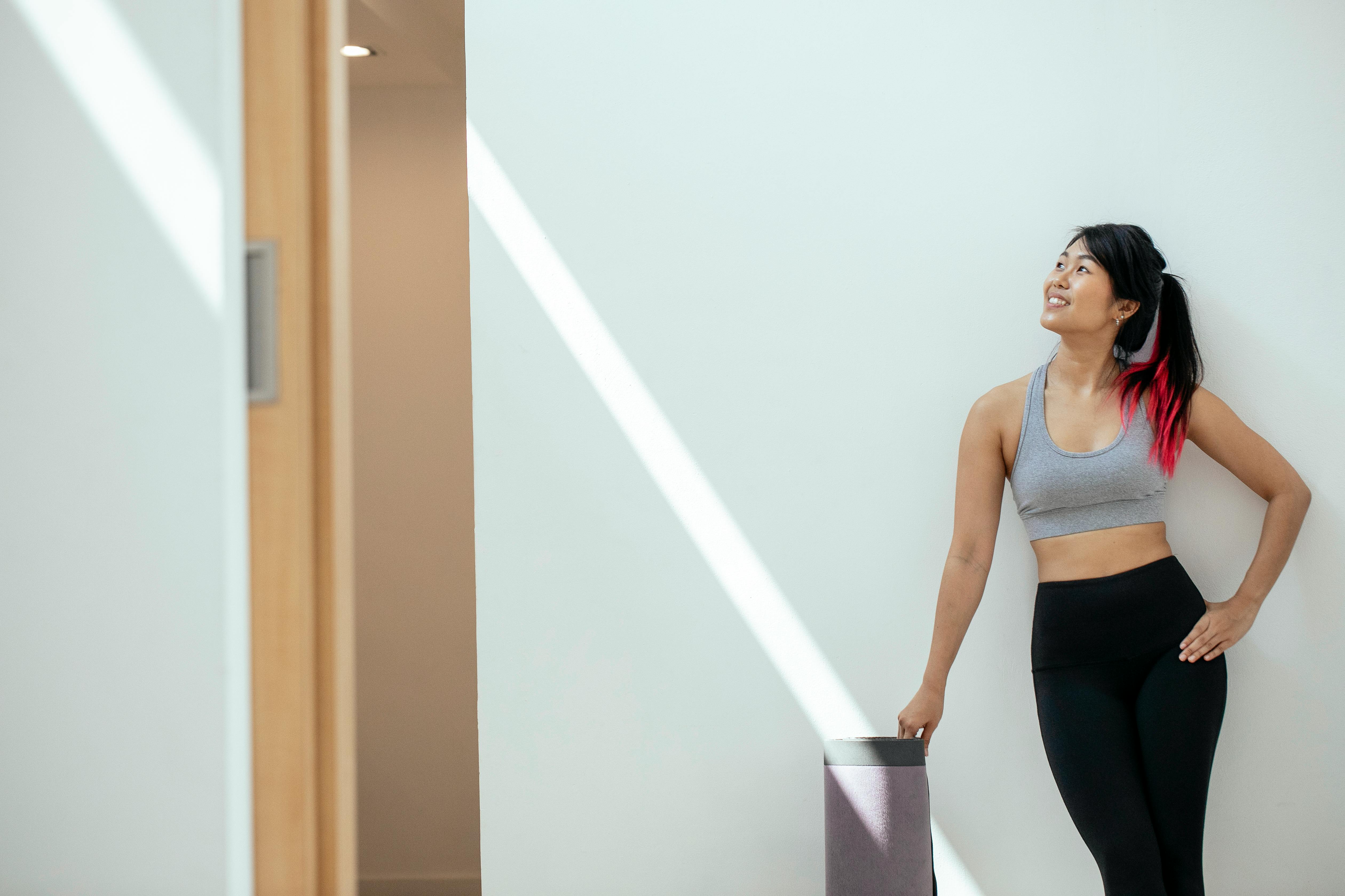 Fit young female in activewear holding mat and standing near white wall while smiling and looking away