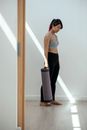 Side view full body of ethnic sportswoman in activewear holding rolled mat while looking down and standing in empty light room