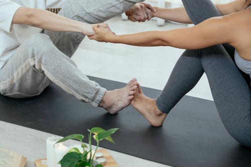 Crop unrecognizable barefoot woman and man sitting together on yoga mat and holding hands while practicing yoga in light apartment