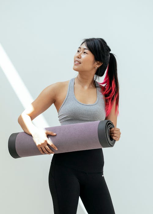 Fit Asian female in activewear with ponytail holding rolled yoga mat while looking away and standing on white background