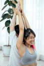 Young Asian female in gray top sitting back to back with male partner and keeping hands raised while practicing yoga in light room