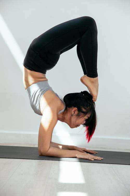 Free Side view of full body flexible Asian female doing Vrischikasana A yoga pose on gray mat in light room Stock Photo