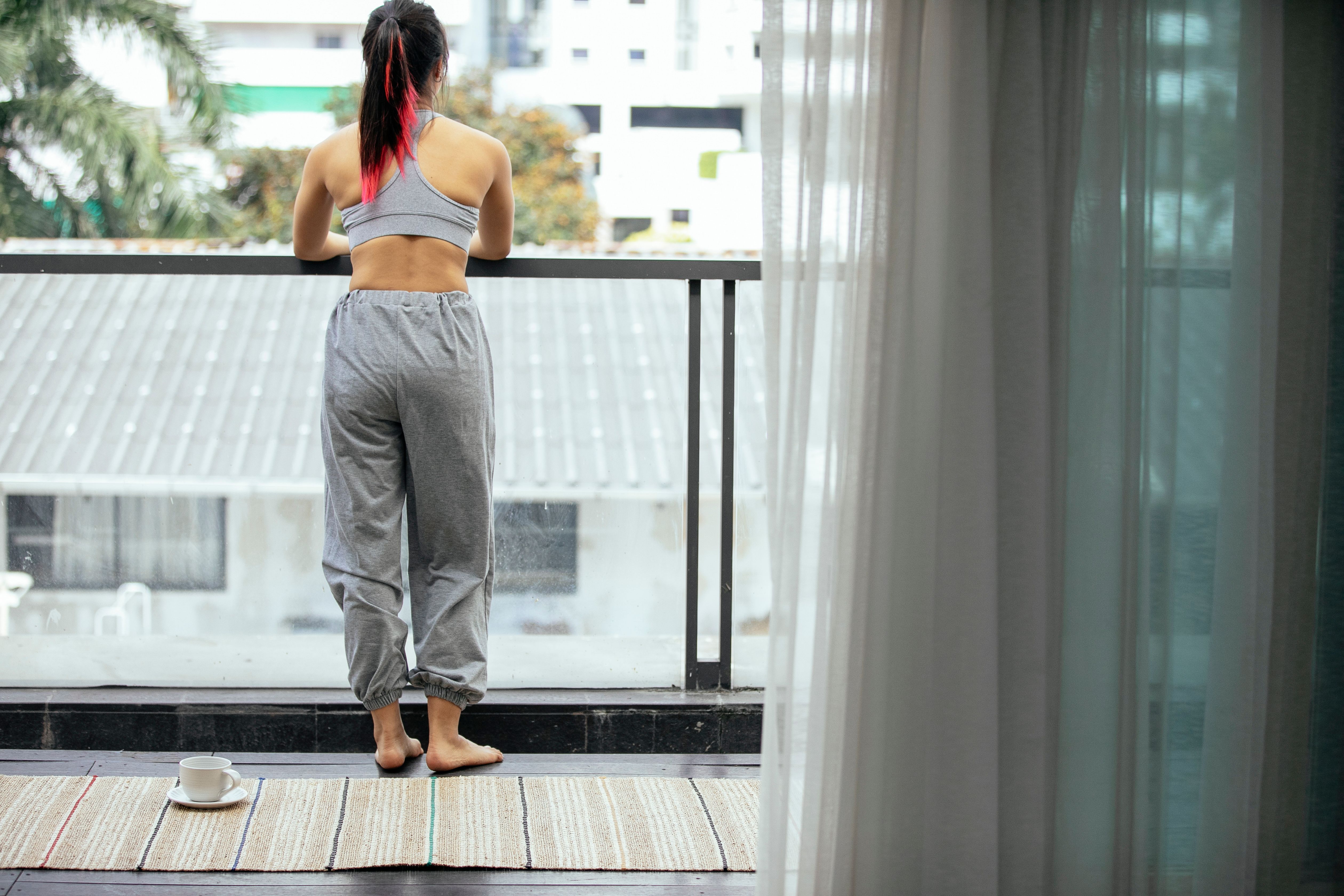 Back view of anonymous peaceful female in activewear standing on terrace with mat and cup of coffee while enjoying morning and observing city