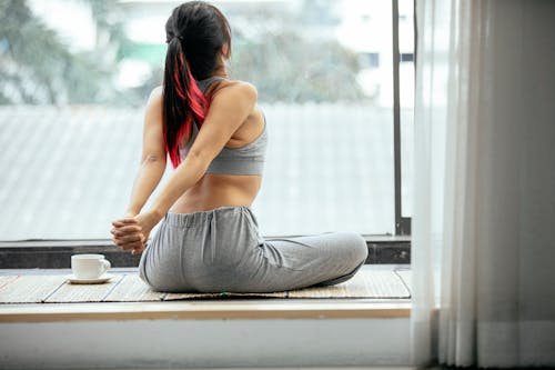 Free Back view of slim unrecognizable female sitting on mat and stretching arms behind back while doing warm up exercise in morning on terrace Stock Photo