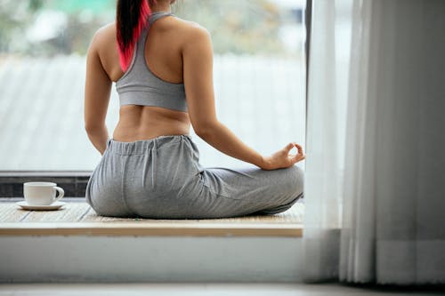 Back view of unrecognizable crop female in sportswear sitting in Lotus pose with mudra hands and practicing yoga on terrace