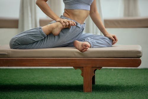 Side view of crop anonymous flexible female in sportswear performing Eka Pada Raja Kapotasana pose on bench during yoga session