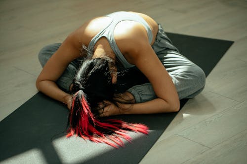 From above full body of anonymous flexible female with long dyed hair bending forward while practicing Baddha Konasana B yoga pose on mat in studio