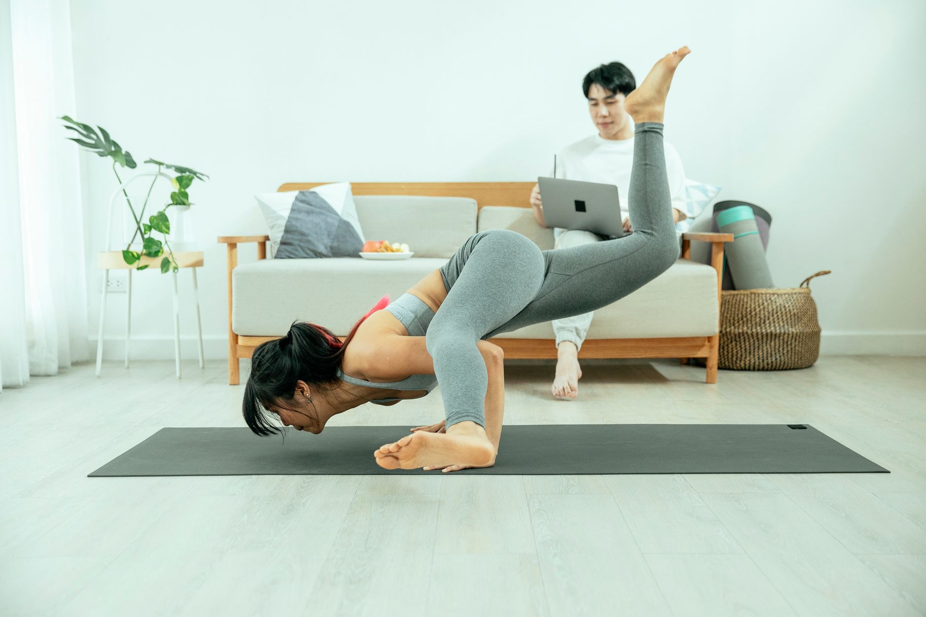Side view of concentrated female practicing yoga asana on mat while Asian male sitting on sofa and using laptop in modern apartment