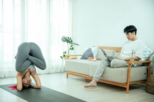 Free Side view of young Asian female doing yoga exercise while man sitting on couch and using laptop in light apartment Stock Photo
