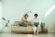 Full body of Asian male using laptop while happy female stretching body on sofa in light living room
