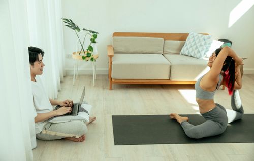 Free High angle of flexible young female in sportswear sitting in yoga pose while Asian male sitting with laptop on floor Stock Photo