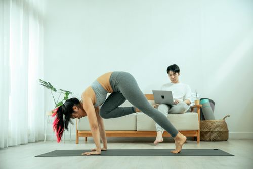 Free Full body of fit female in sportswear doing yoga on mat while Asian male using laptop on sofa in living room Stock Photo