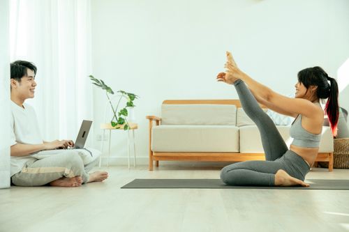 Free Side view of smiling female stretching leg on mat while Asian male using laptop in modern apartment Stock Photo