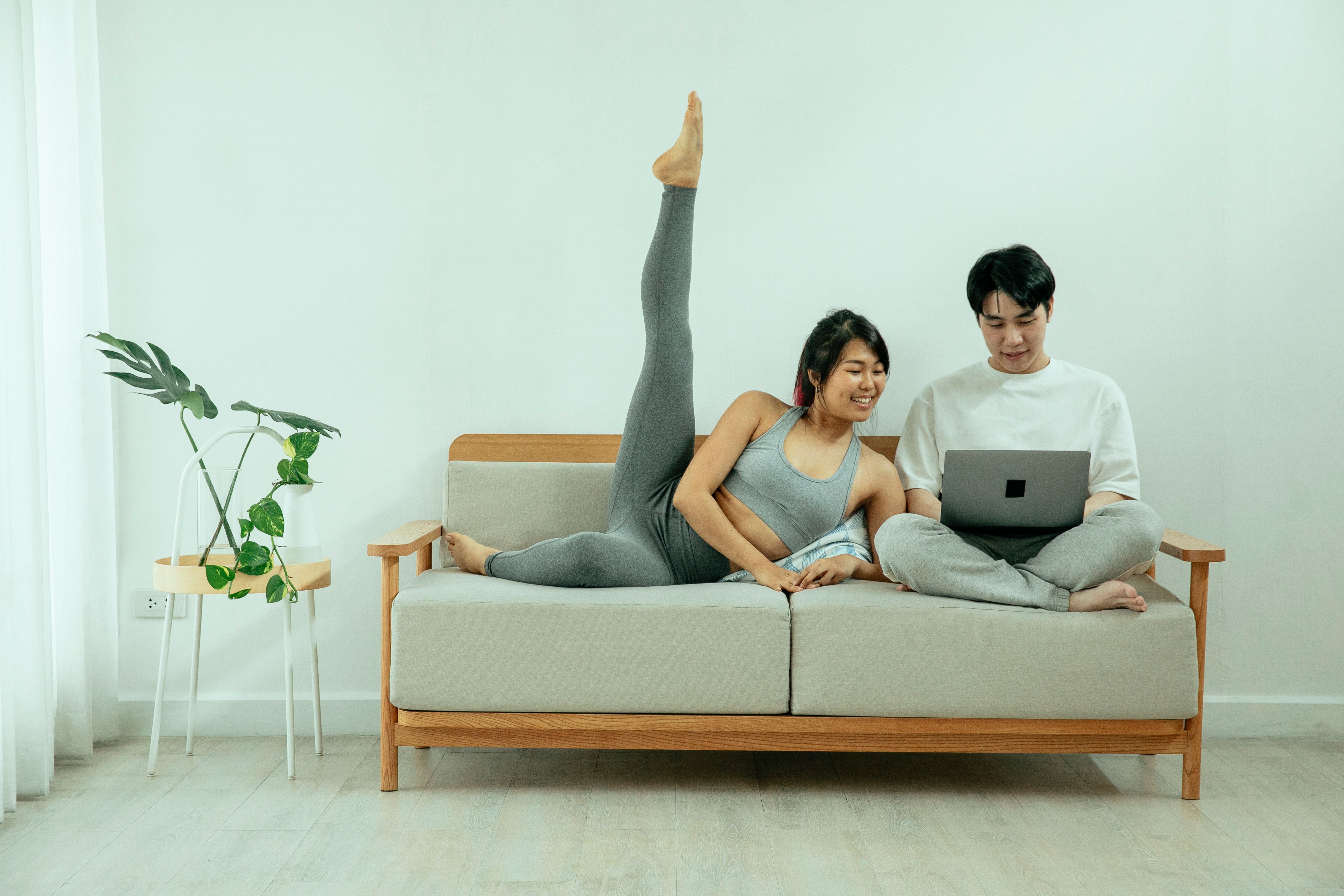 Full body of positive Asian female stretching legs on sofa while male using laptop in modern apartment