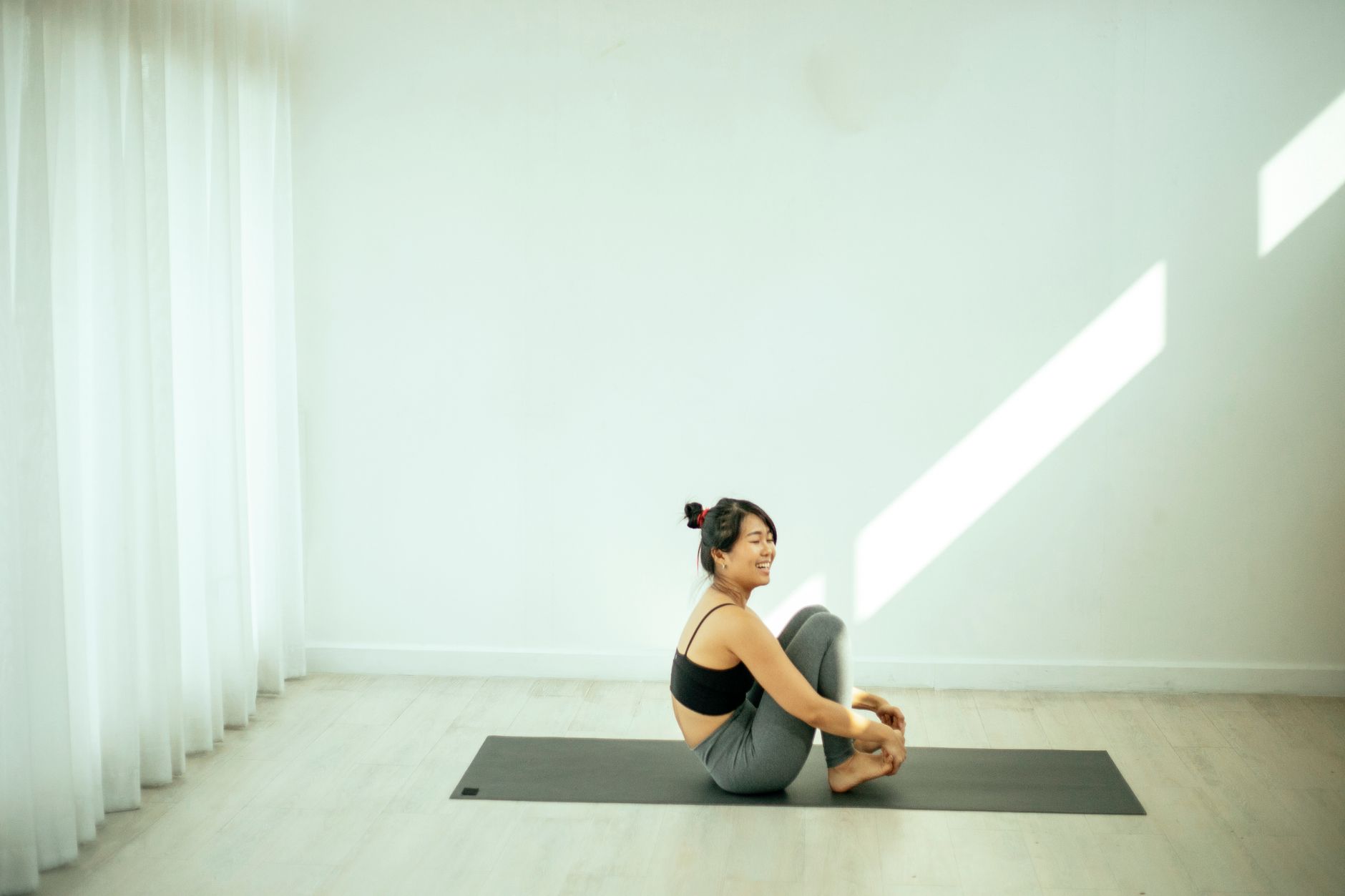 Side view full body of positive Asian female in sportswear sitting with bent legs on yoga mat while looking away