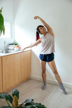 Full body of positive Asian female doing warm up exercise with raised arm while standing near kitchen counter at home