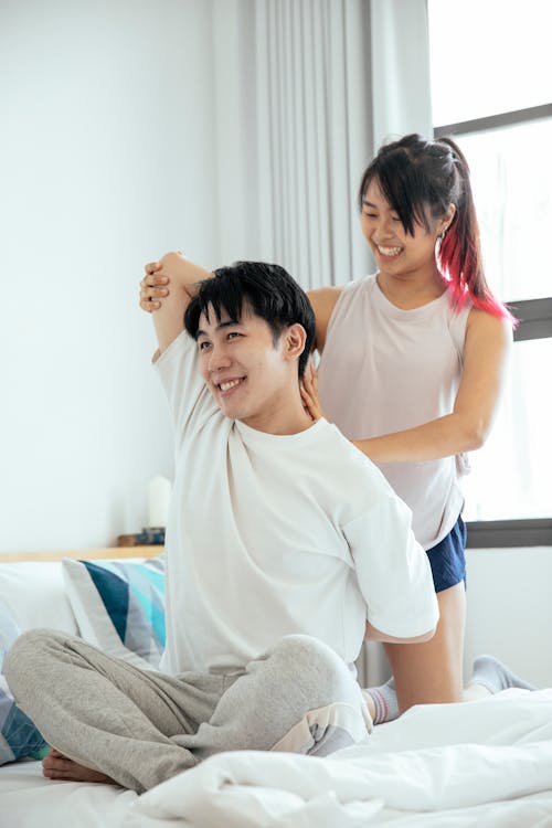 Free Positive Asian girl trying optimistic Asian man to do cow face asana while sitting on comfortable bed in bedroom during yoga lesson Stock Photo