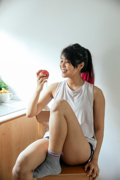 Free A young woman sitting indoors, joyfully holding an apple with natural lighting. Stock Photo