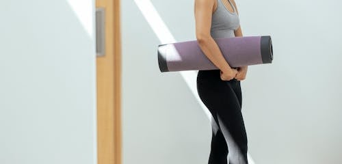 Crop sportive woman with rolled yoga mat standing in studio
