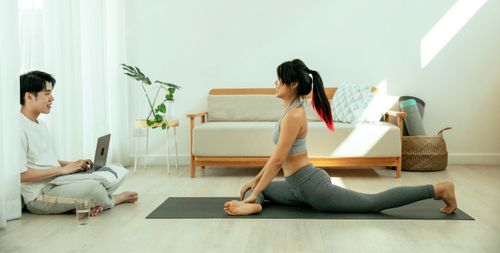 Free Side view of concentrated barefoot female in sportswear performing Half Pigeon pose on mat during yoga session and smiling male sitting on floor crossed legs with netbook Stock Photo
