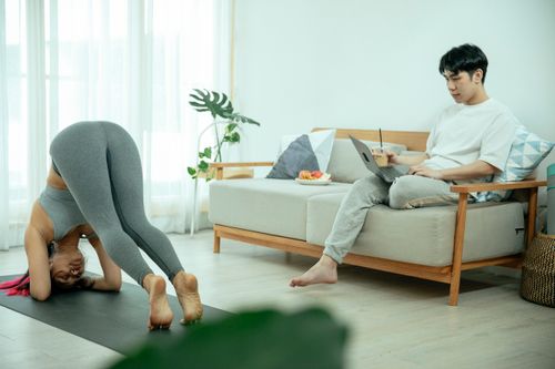 Free A woman exercises while a man relaxes with a laptop in a cozy, modern living room. Stock Photo