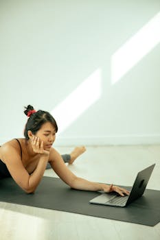 Side view of concentrated ethnic female in activewear stretching legs on mat on floor while browsing netbook in light room and looking at screen resting head on hand