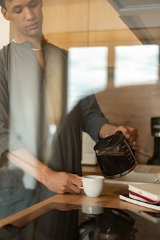 Young man pours coffee into a ceramic mug in a cozy indoor kitchen, captured in a vertical frame.