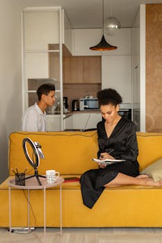 Woman in a black dress writing in a notebook on a yellow sofa indoors during daytime.