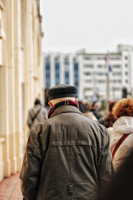 Back view of unrecognizable senior male in outerwear and cap walking on street in crowd on blurred background