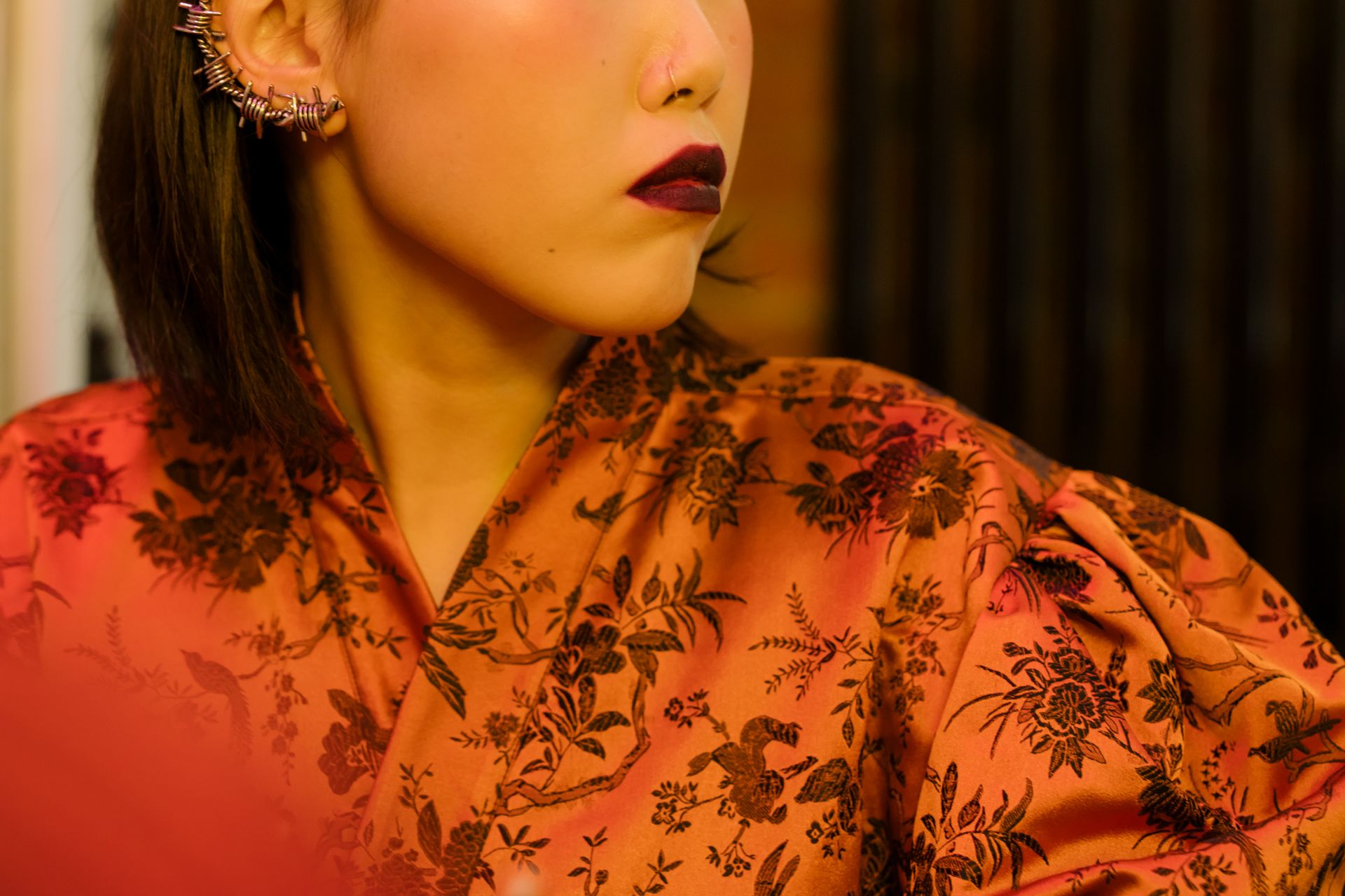 Fashionable woman in profile wearing a floral brocade dress with bold makeup and ear cuff in an indoor setting.