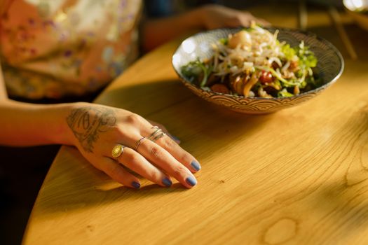 Close-up of a tattooed hand with rings, holding a bowl of noodles on a wooden table, sunlit atmosphere.