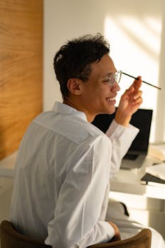 A cheerful man in a white shirt sitting at a desk with a laptop, enjoying the warm indoor setting.