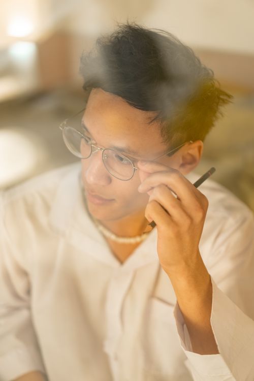 Free A young man wearing glasses holds a pen thoughtfully, captured in soft warm tones. Stock Photo