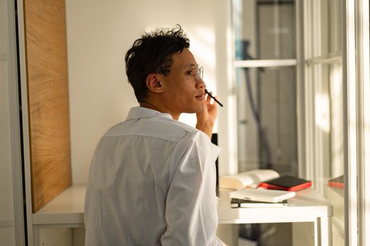 Man in white shirt gazing out window pensively, surrounded by books in modern office.
