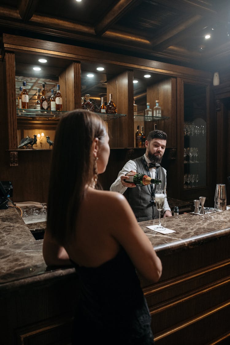 A Bartender Behind Bar Counter Pouring Wine In A Glass Near A Person