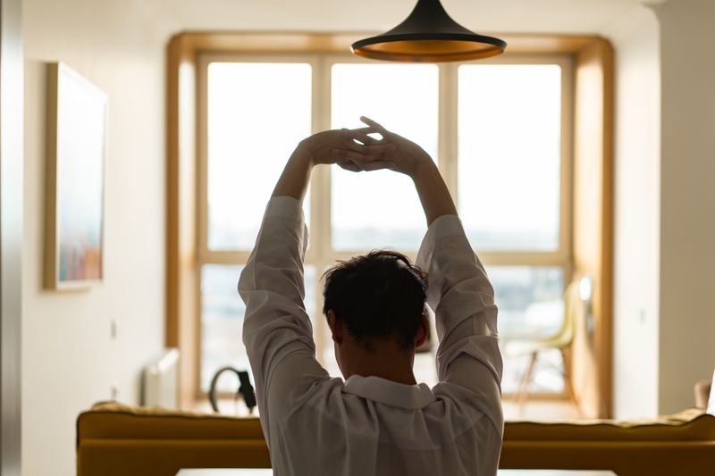 Back view of a man stretching inside a room with a sunny window backdrop