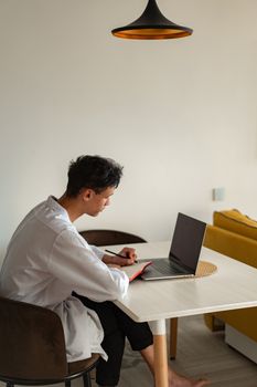 Young man in white shirt working on laptop while taking notes in modern home setting.