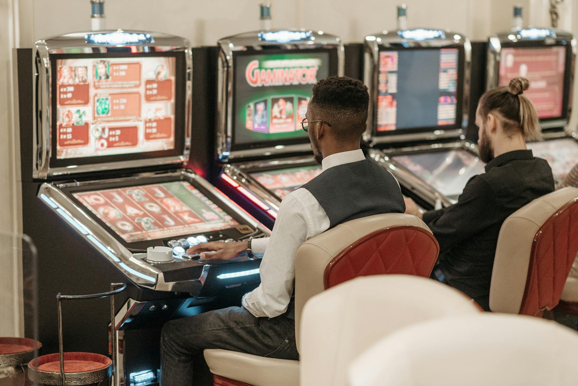 Two men engaged in gaming at a casino, seated at slot machines indoors.