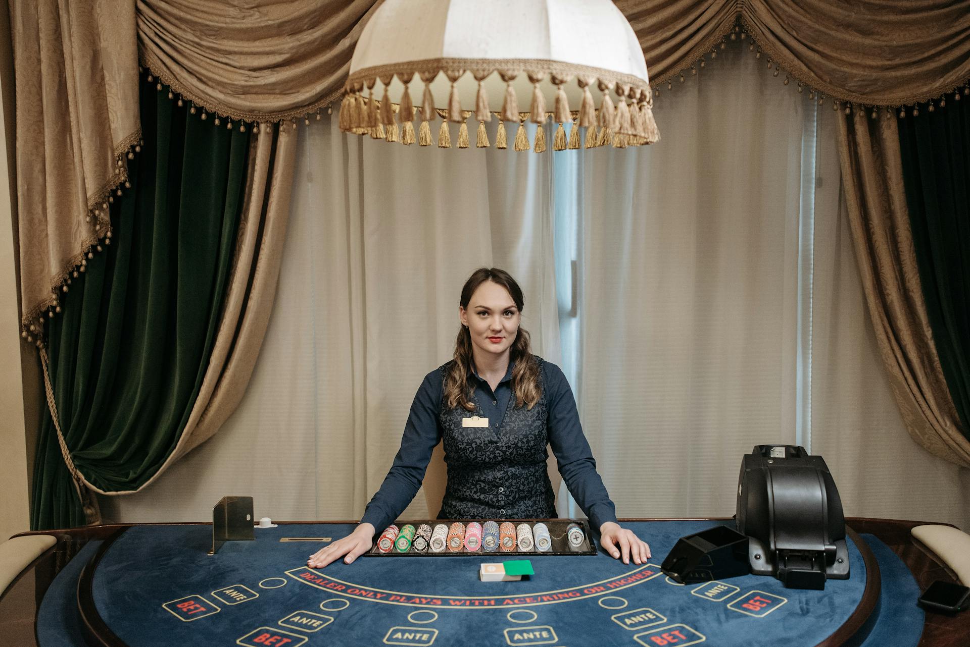 A smiling female casino dealer at a gaming table surrounded by chips and cards indoors.