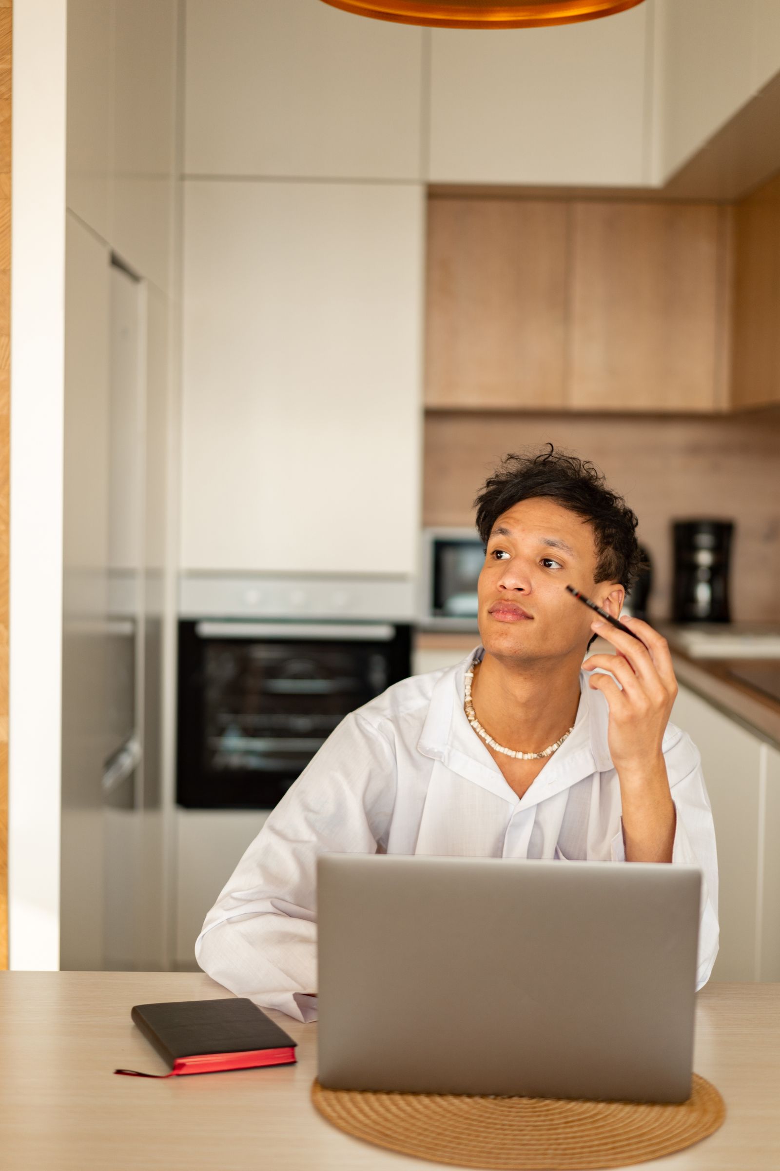 A young man in a white shirt holding a pen beside his laptop and notebook at home