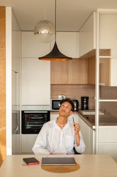 Man in a white shirt sits at a table, looking up, in a modern kitchen setting.