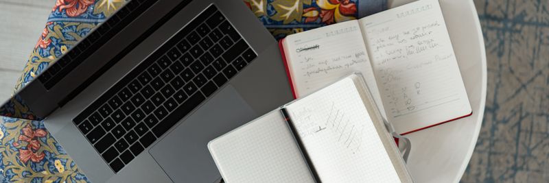 Overhead shot of a laptop with notebooks and a pencil on a patterned cloth.