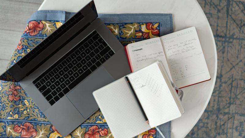 Overhead shot of a laptop with notebooks and a pencil on a patterned cloth.