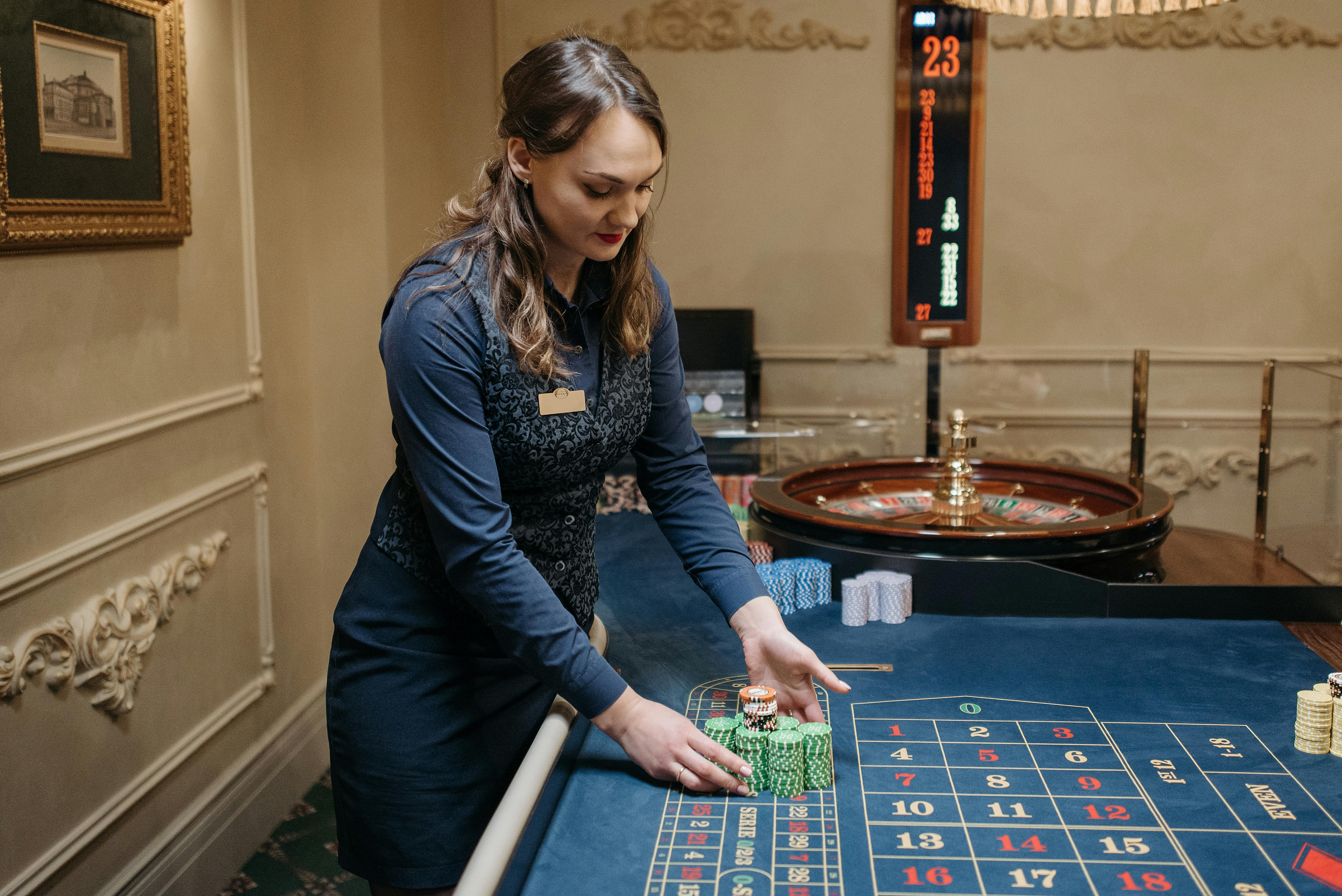 Female casino dealer arranging chips on a roulette table in an elegant gaming room.