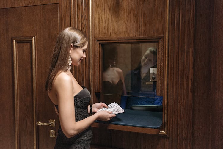 A Woman Holding A Large Amount Of Money While Standing On A Cashier Window