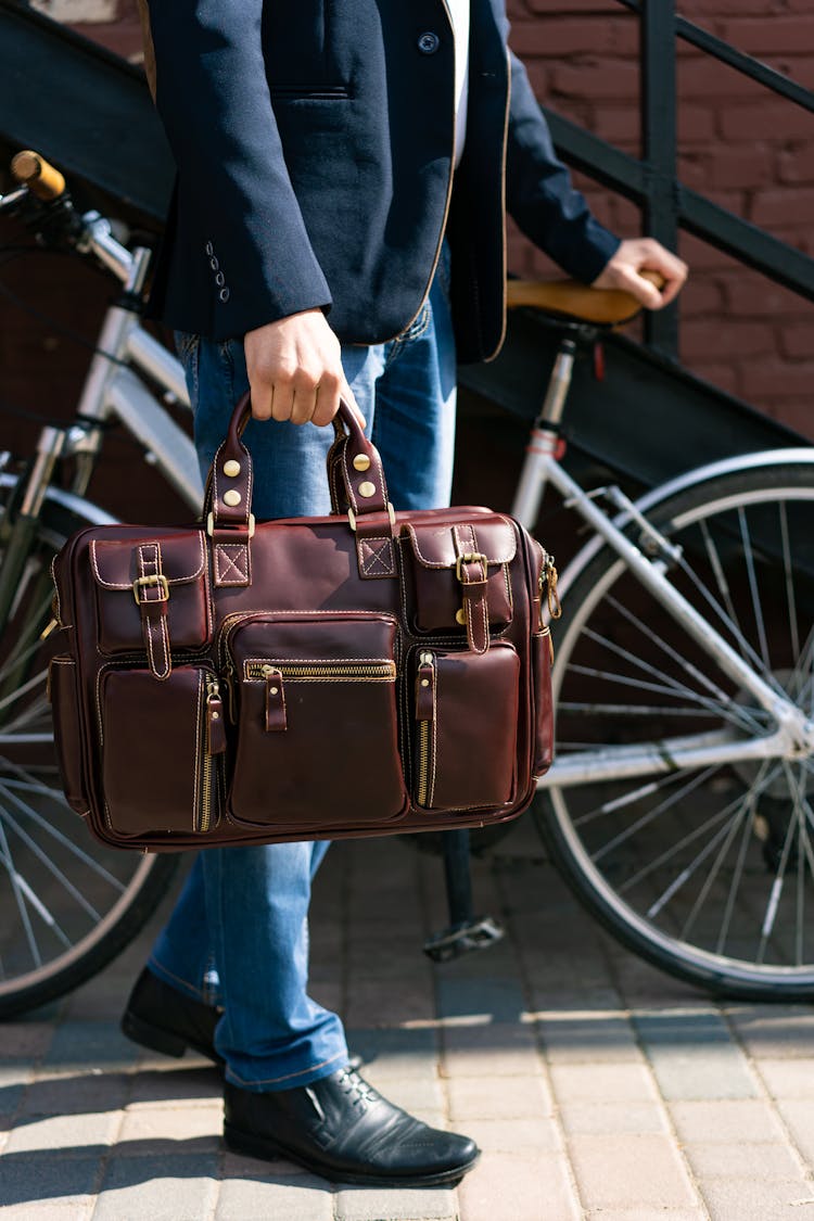 A Man Standing Beside A Bicycle While Holding A Brown Leather Briefcase Bag