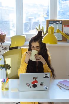 Woman sitting at desk with laptop, enjoying coffee, in a home office setting filled with light and plants.