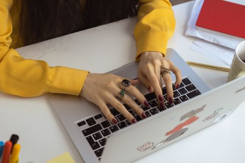 Free Close-up of a woman typing on a laptop, showcasing red nails and rings, wearing a yellow long sleeve indoors. Stock Photo