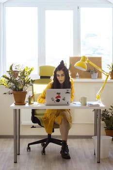 Hispanic woman in yellow jacket working from a home office desk with plants and a desk lamp.